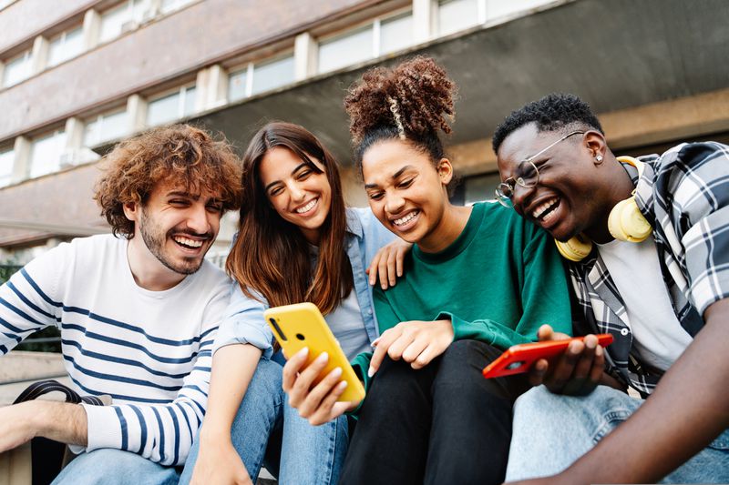 Group of smiling young adults sitting together outside, using mobile phones.
