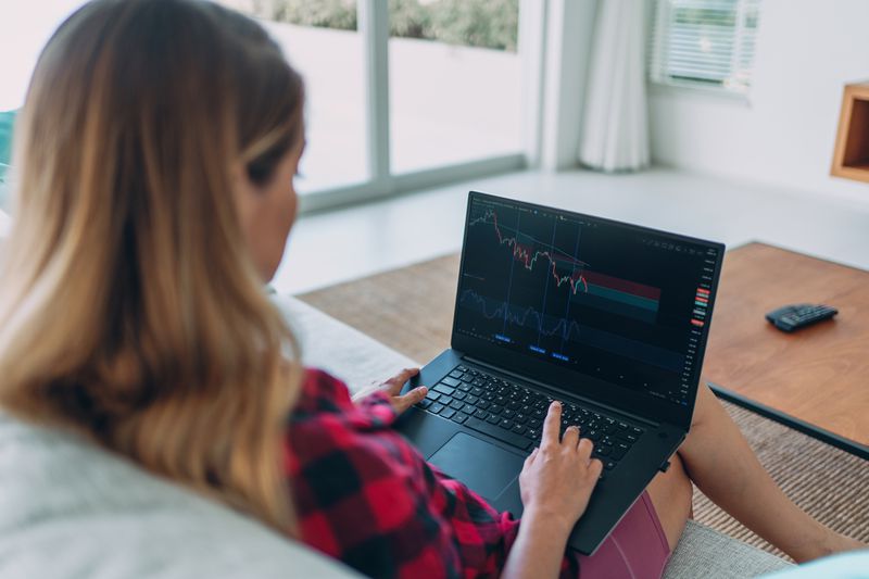 A woman with her back to the camera sits on a couch in a living room, using a laptop displaying financial charts.