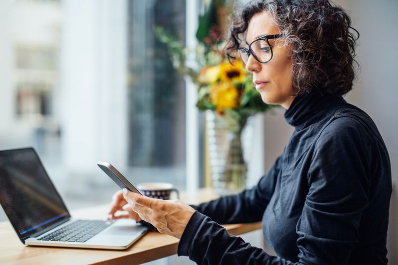 businesswoman sitting at cafe looking at her cell phone while working on laptop computer. Woman reading text message at coffee shop.