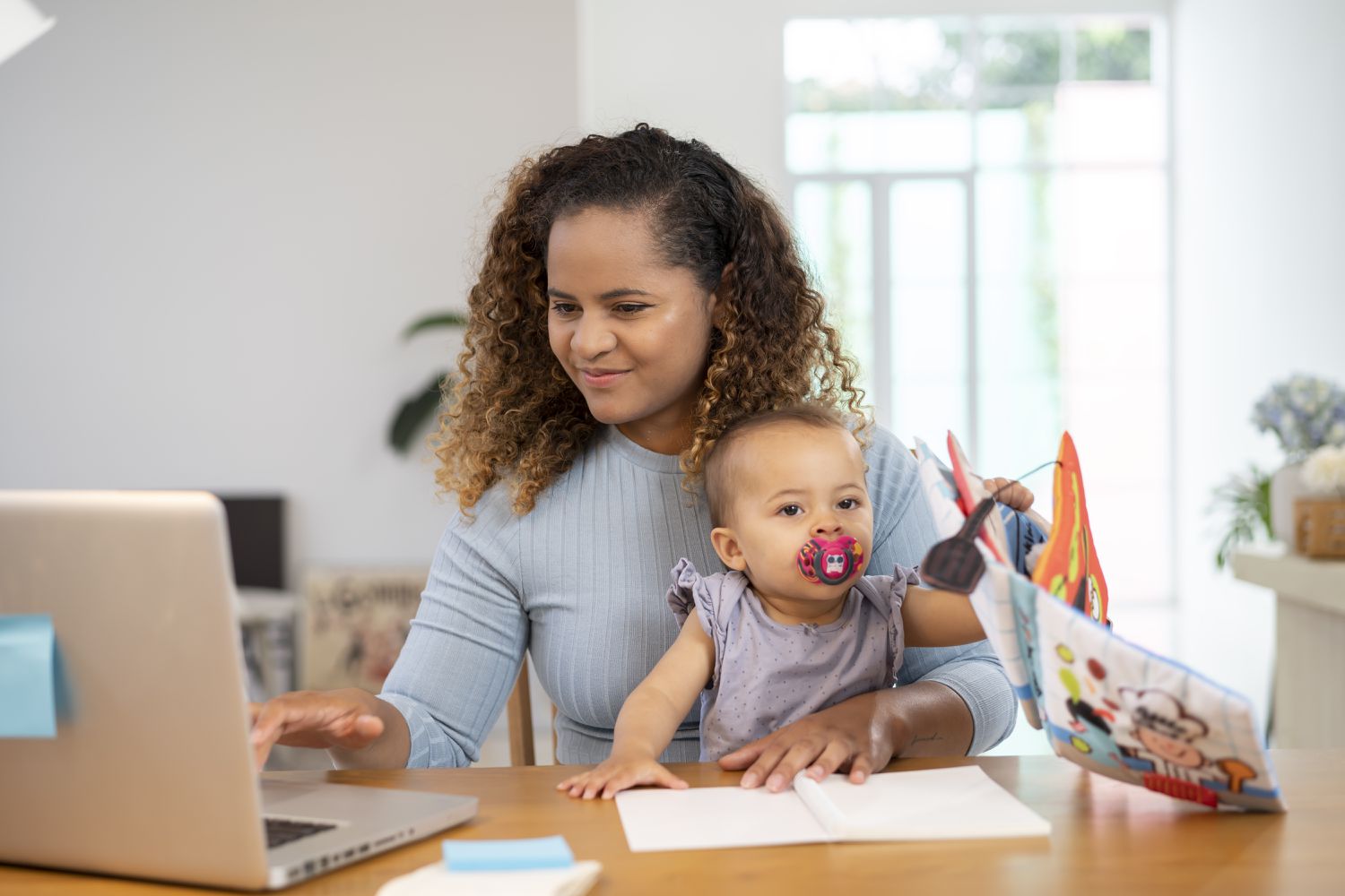 Young mom sitting at dining room table with baby on her lap, checking something on her laptop