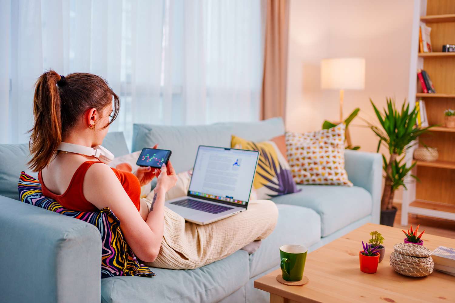 Woman sitting on a couch looking at charts on her cell phone with her laptop on her lap.
