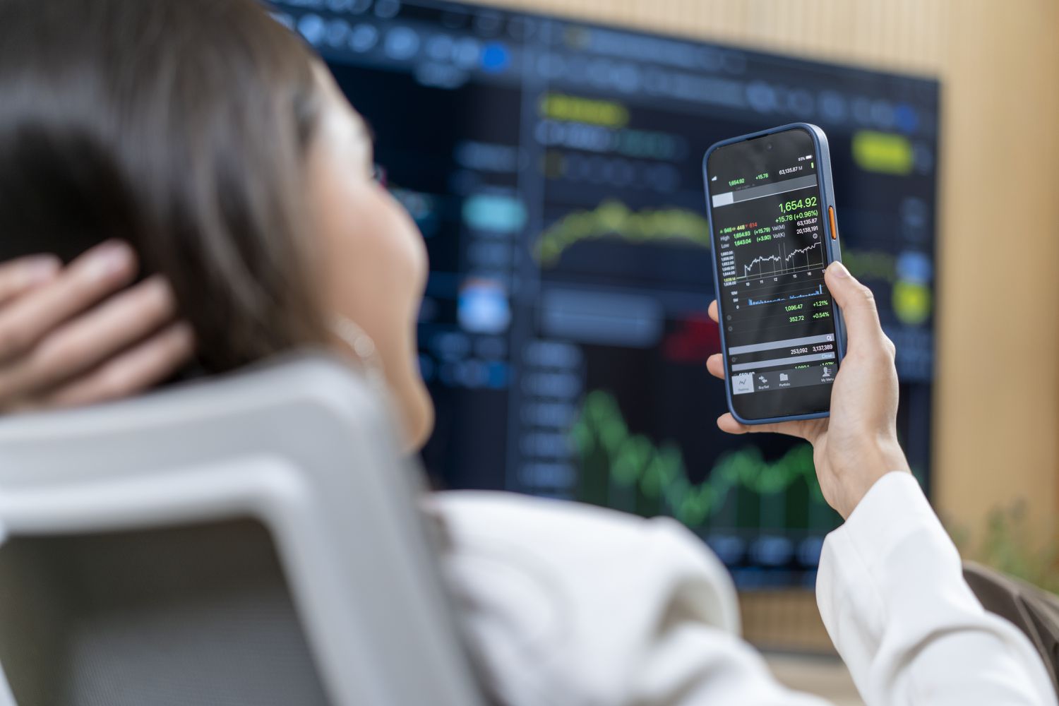 Woman looking at a stock chart on a smartphone with another larger screen with stock charts on it in the background
