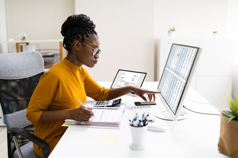Woman working at desk writing in a binder with a laptop and desktop on her table checking financial information