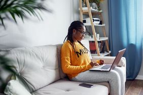 Young Black woman looking at laptop while sitting on sofa.
