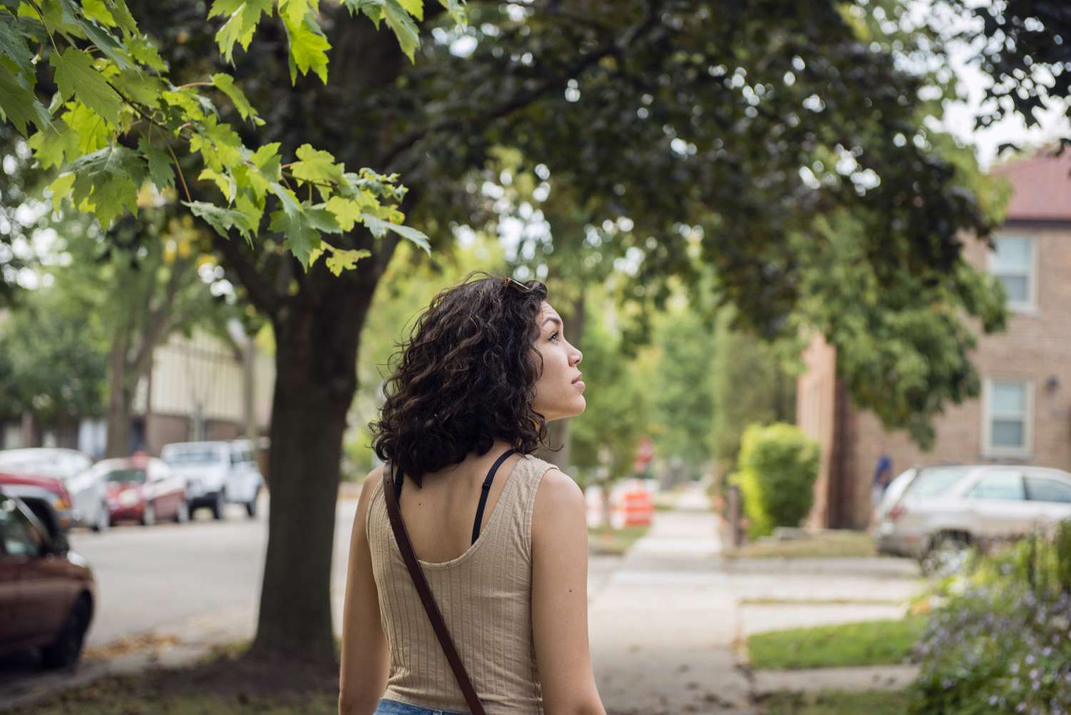 A woman with dark hair and a beige tank top walks down the street looking at houses