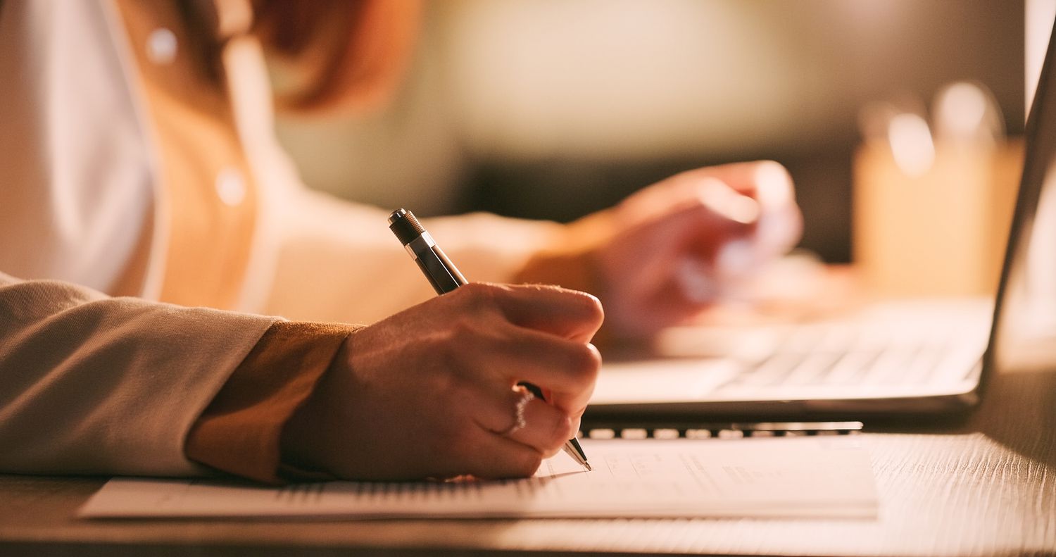 Hands of woman holding a pen and writing on a desk with a laptop next to her