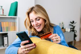 Similing young woman holding a phone and a credit card while sitting on a yellow couch. 