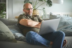 Man in his 50s or 60s sitting on his sofa and looking with concern at his laptop
