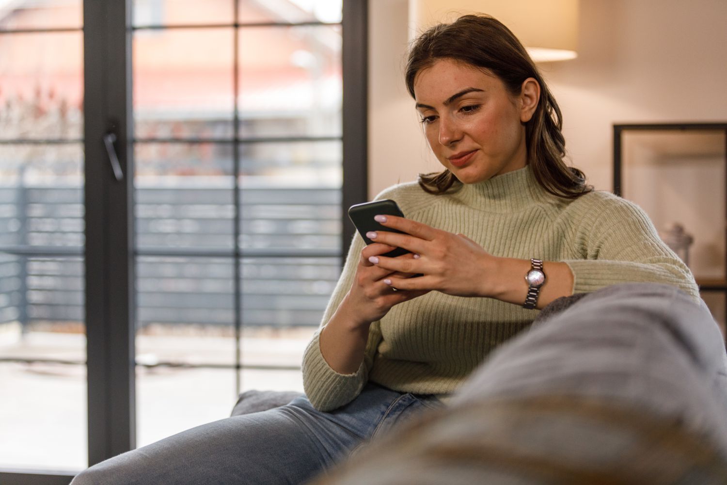 Young woman sitting in her living room and looking seriously at something on her smartphone
