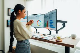 Woman standing win front of a desk with multiple monitors showing charts and other data.
