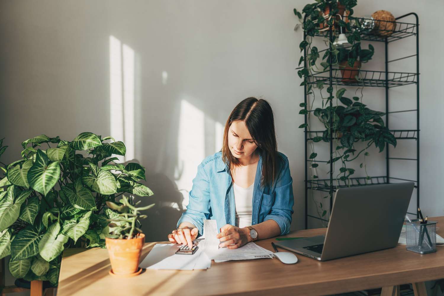 Woman using a calculator to calculate finances at a desk surrounded by greenery and natural sunlight.