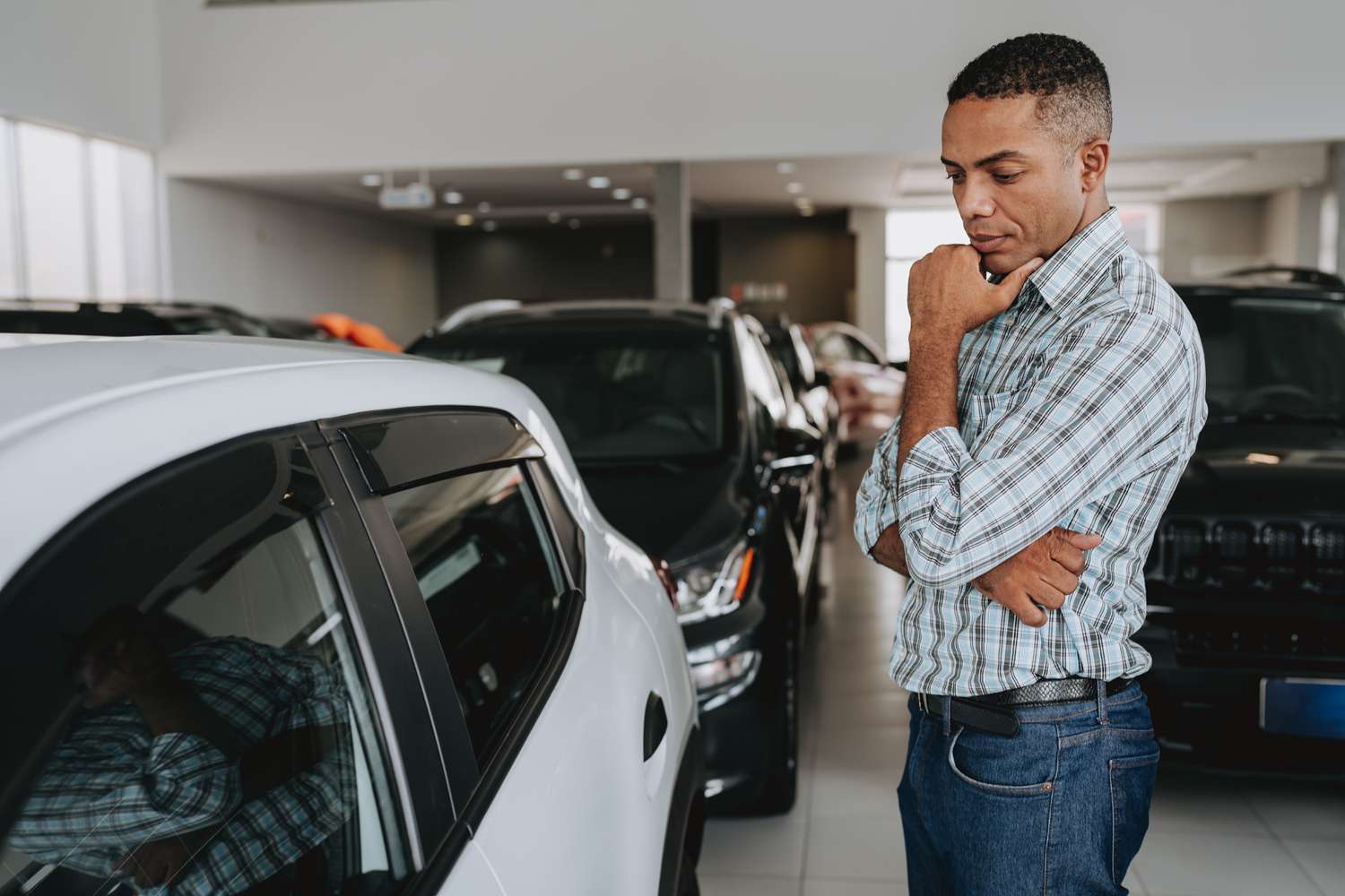 A car buyer inside a car dealership standing in front of a new vehicle and contemplating the cost.