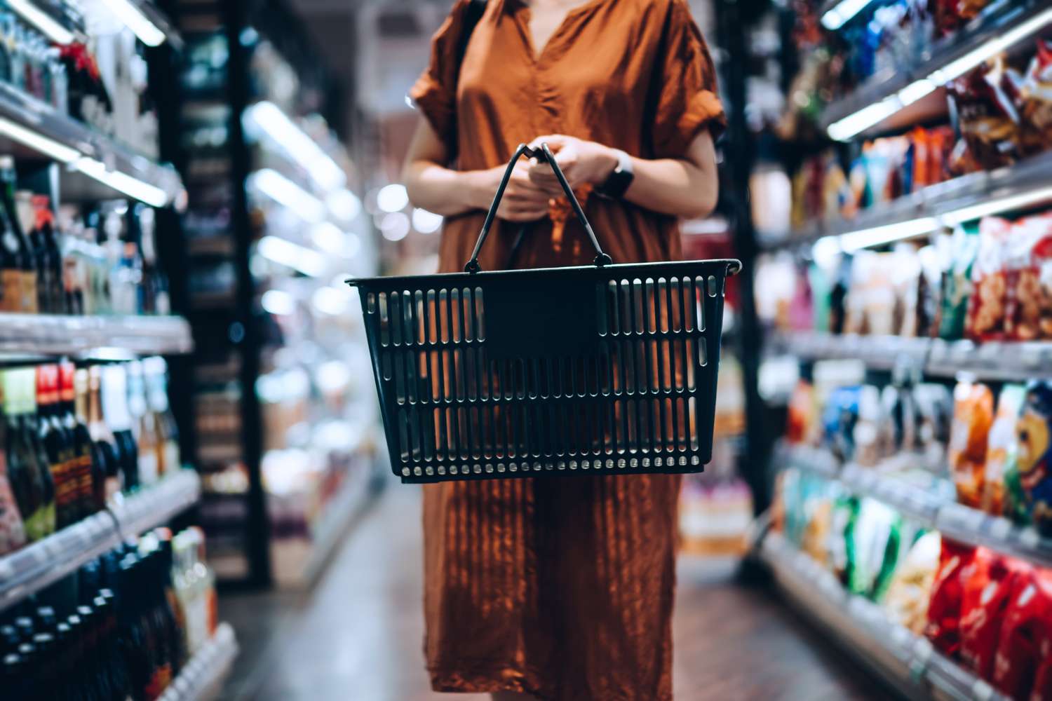 A shopper carries an empty shopping basket down a grocery store aisle lined with products.