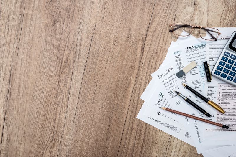 Tax forms on wooden table with notepad, pen, pencil, glasses, and calculator