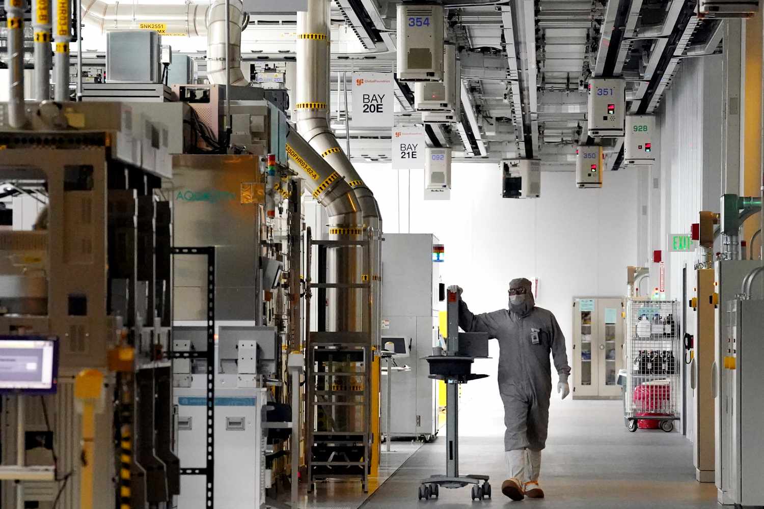 An employee wearing a cleanroom suit walks beneath Automated Material Handling Systems (AMHS) vehicle robots moving along tracks on the ceiling inside the GlobalFoundries semiconductor manufacturing facility in Malta, New York, US, on Tuesday, June 18, 2024.