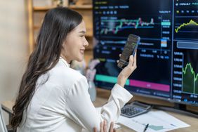 Woman sitting at a desk in front of a large monitor showing multiple chats while looking at a phone.