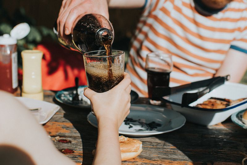 A child holds a glass out toward another person, who fills it with a fizzy drink.