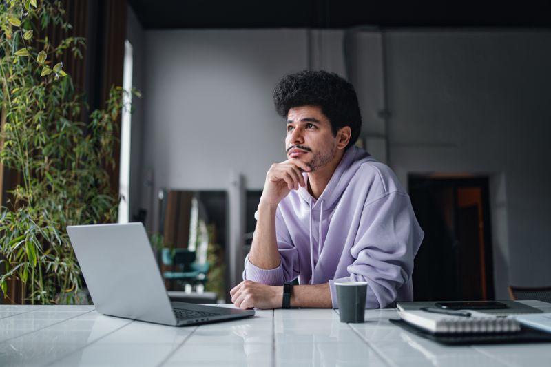 A man sits with a laptop at a table, looking into the distance and pondering.