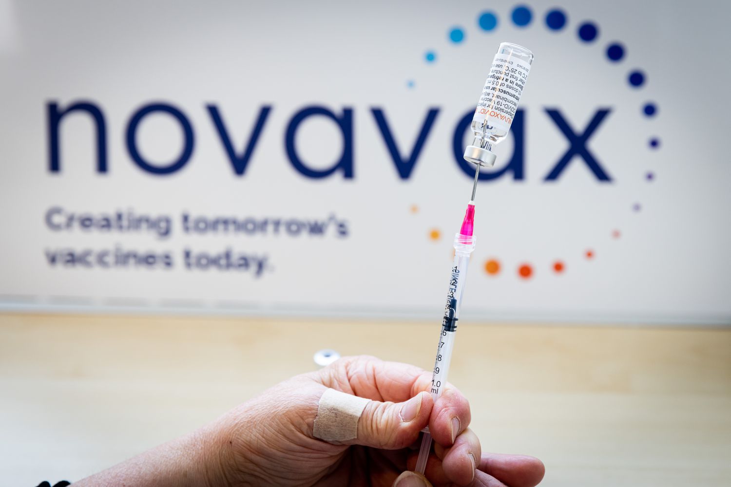  A close-up of a person's hand preparing a dose of a Novavax vaccine, with the company's logo in the background.