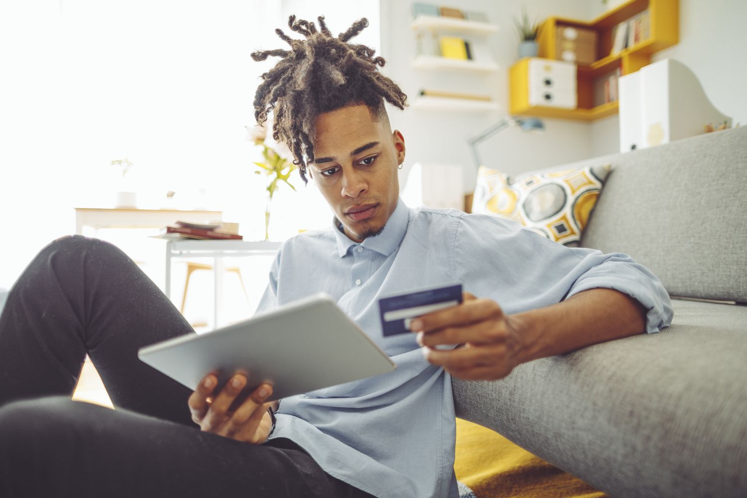 A young man looks at his credit card while holding a tablet. 
