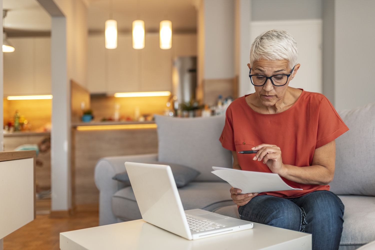 A woman in an orange shirt and jeans sits on her couch looking at a document with a laptop open in front of her on the coffee table.