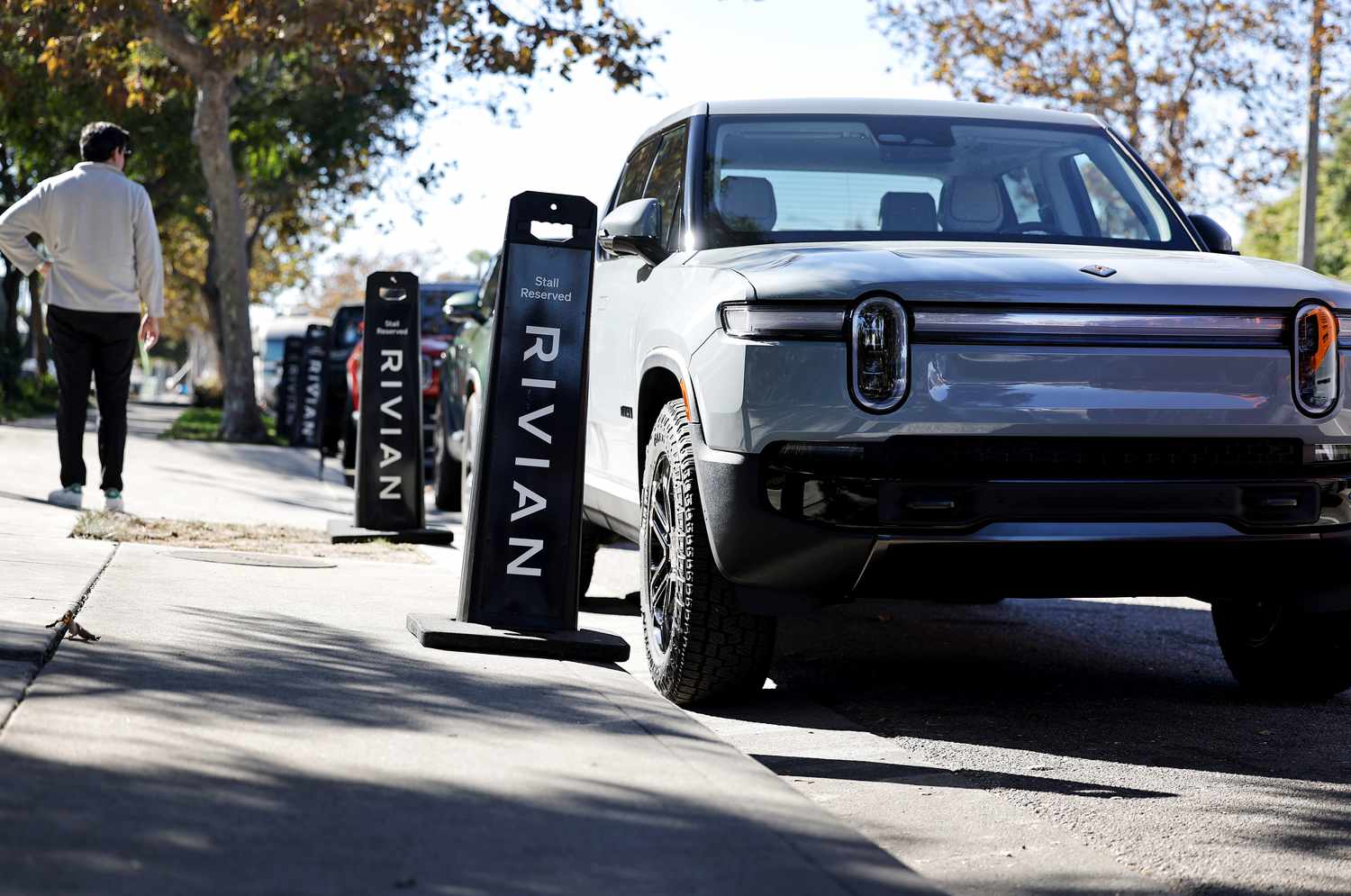A person stands on the sidewalk to the left of several Rivian electric vehicles parked along the street.