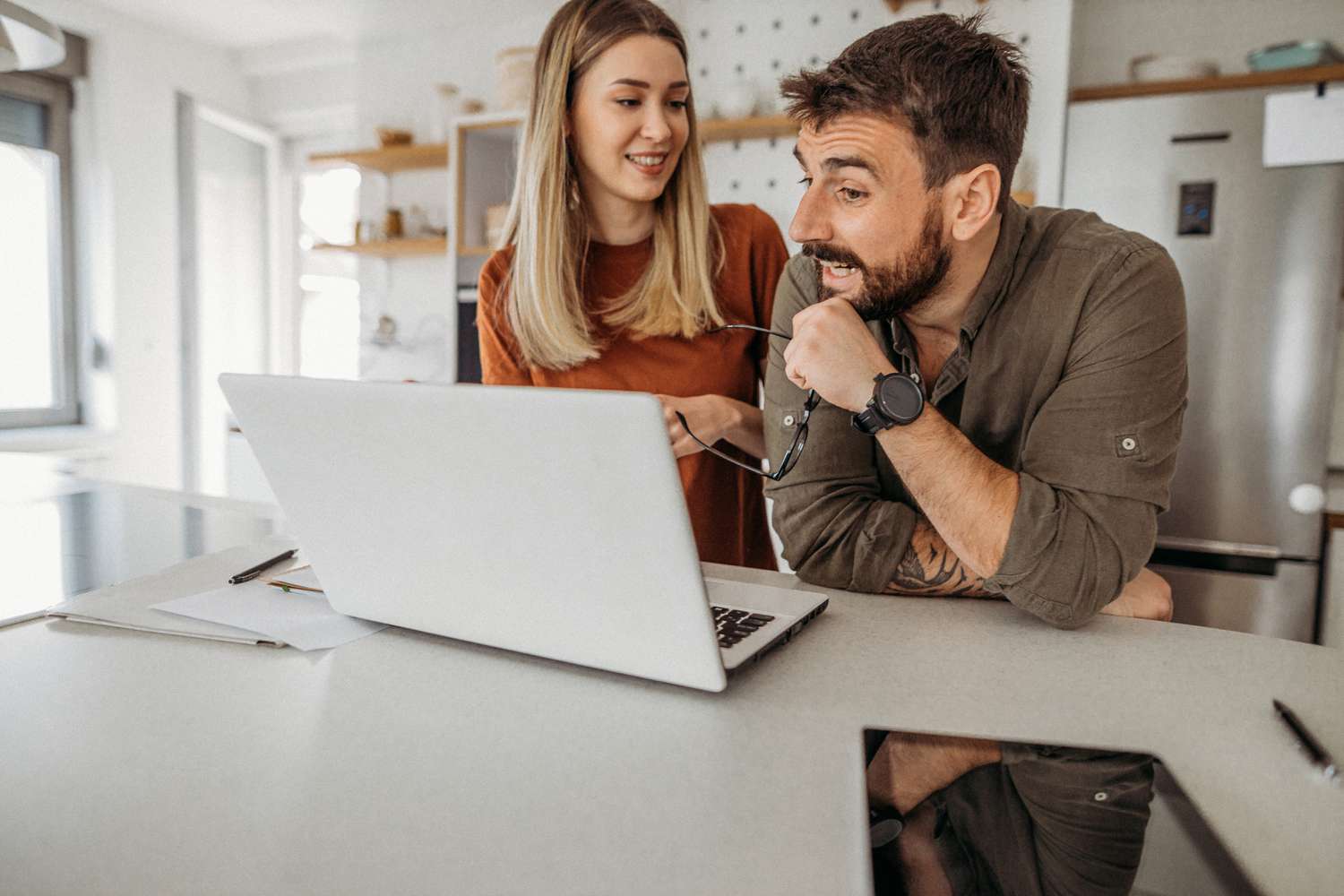 A happy couple stand at their kitchen table, looking at a laptop.