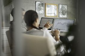 Two people sitting infront of computer monitors showing stock charts and having a discussion.