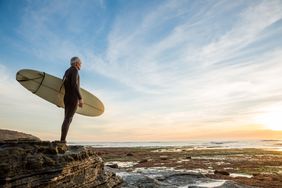 Senior retired surfer man looking at waves and holding a surfboard at Sunset Cliffs, San Diego, California.