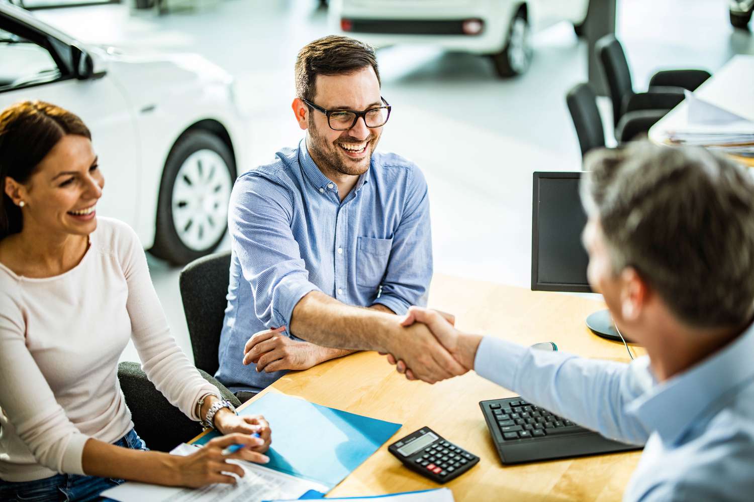 A happy couple buy a car in a showroom, shaking hands with a salesperson.