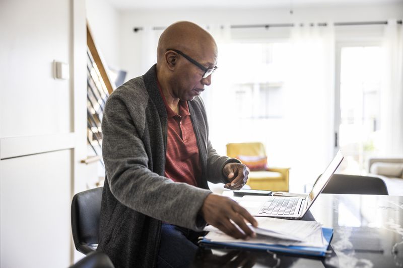 A middle-aged man wearing glasses and a cardigan sits at a table, working on his laptop while handling paperwork. Natural light streams through large windows with sheer white curtains.