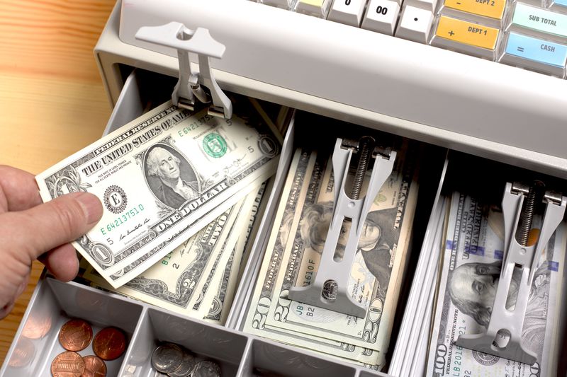 close up shot of the open drawer of a cash till, full of American dollars, with a person putting a $1 bill in.