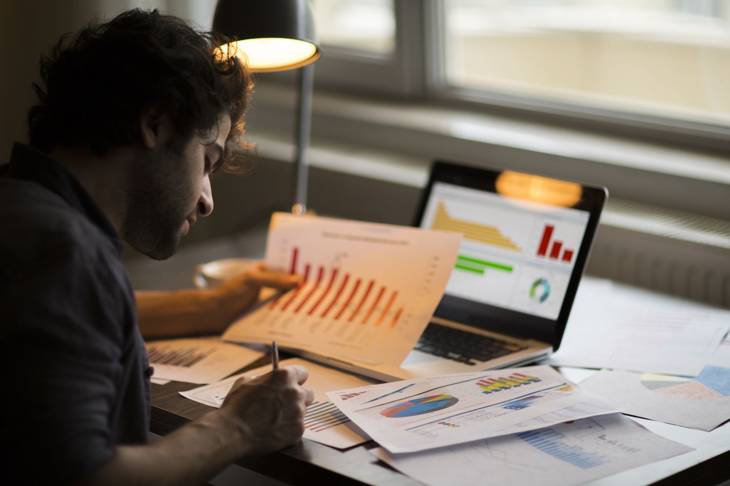 Person writing with a paper with bar graphs in hand and a computer with charts nearby.