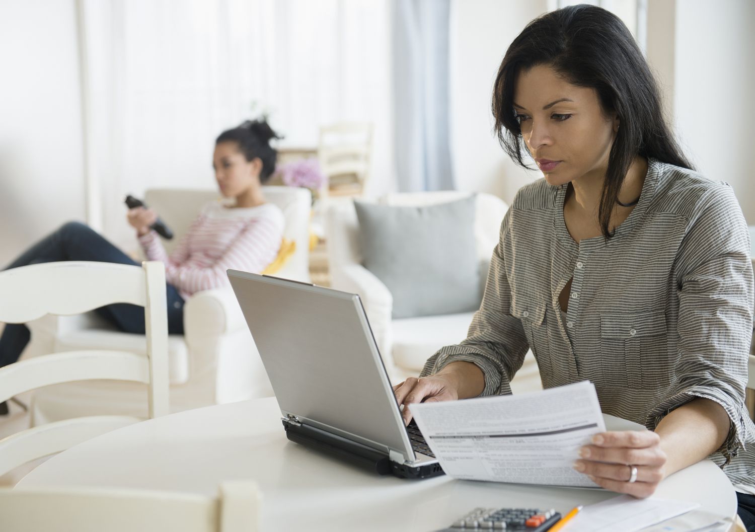 A woman seated at a kitchen table holds a document and peers at a laptop screen. In the background a young woman relaxes in an easy, television remote control in hand.