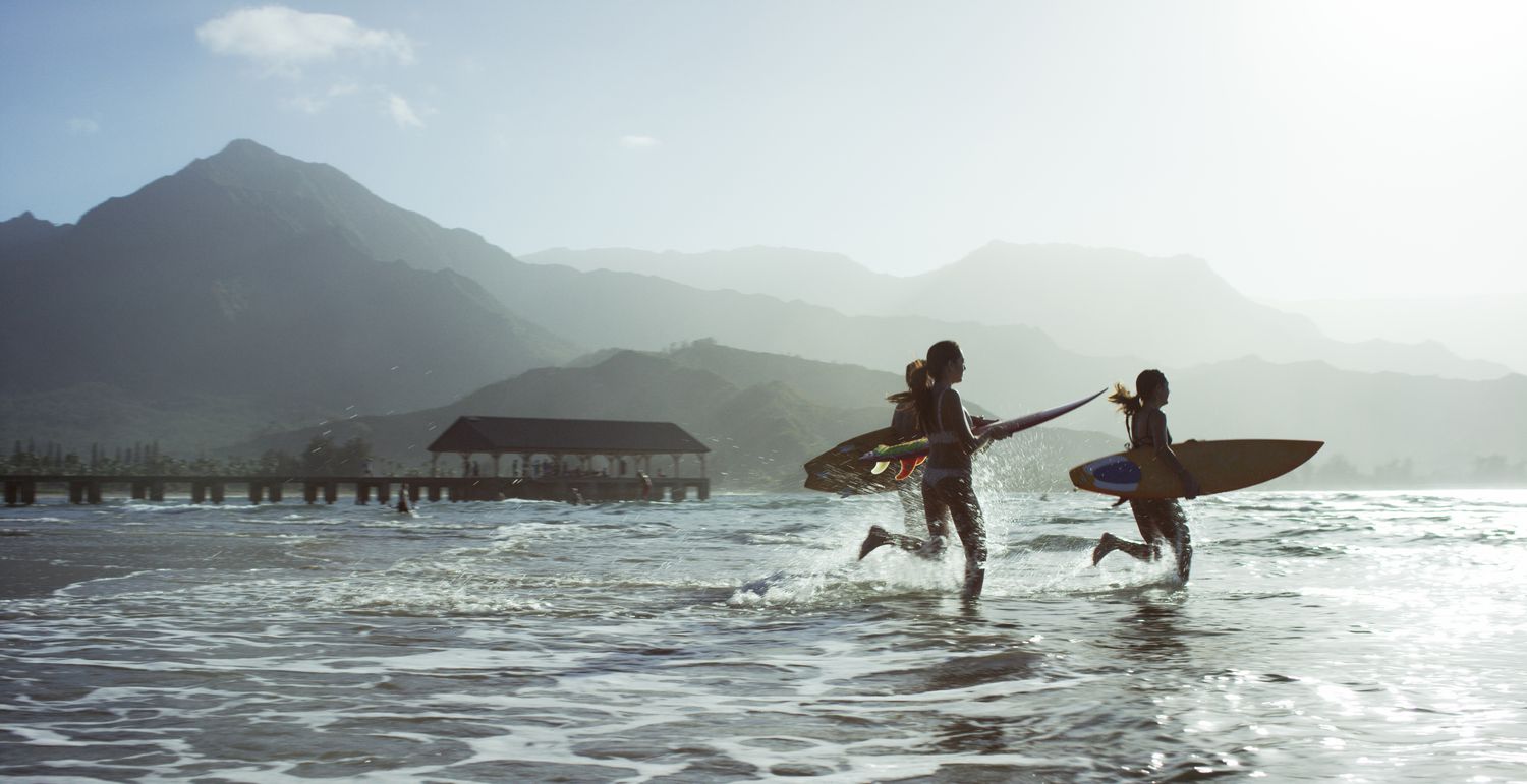 People running on a beach with surfboards with mountains in the background.