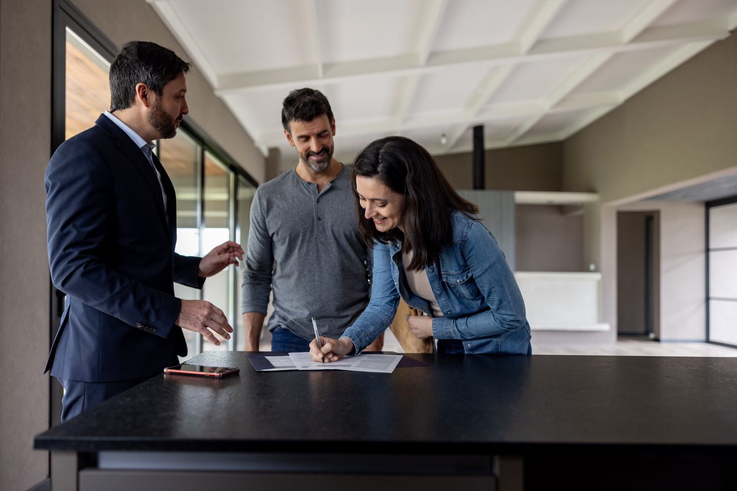 A man and a woman sign documents in an empty house beside a realtor