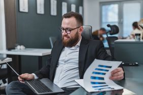 Man in office looking at a laptop while holding document displaying bar charts. 