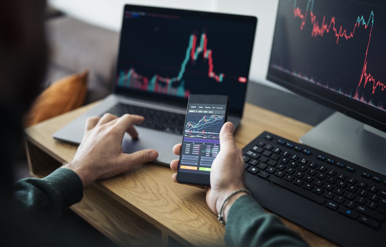 A man holds a cell phone displaying financial charts while sitting in front of two computers at a desk.