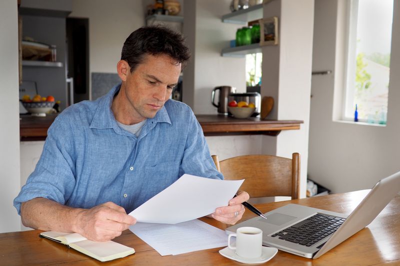 Man in his 40s sitting in his kitchen and looking at financial documents and a laptop
