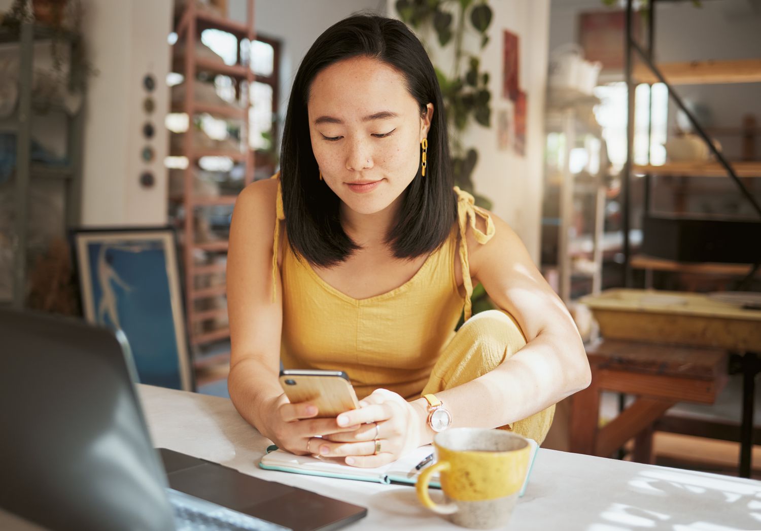 A woman in a yellow outfit sits at a desk with a computer and coffee looking at her smartphone