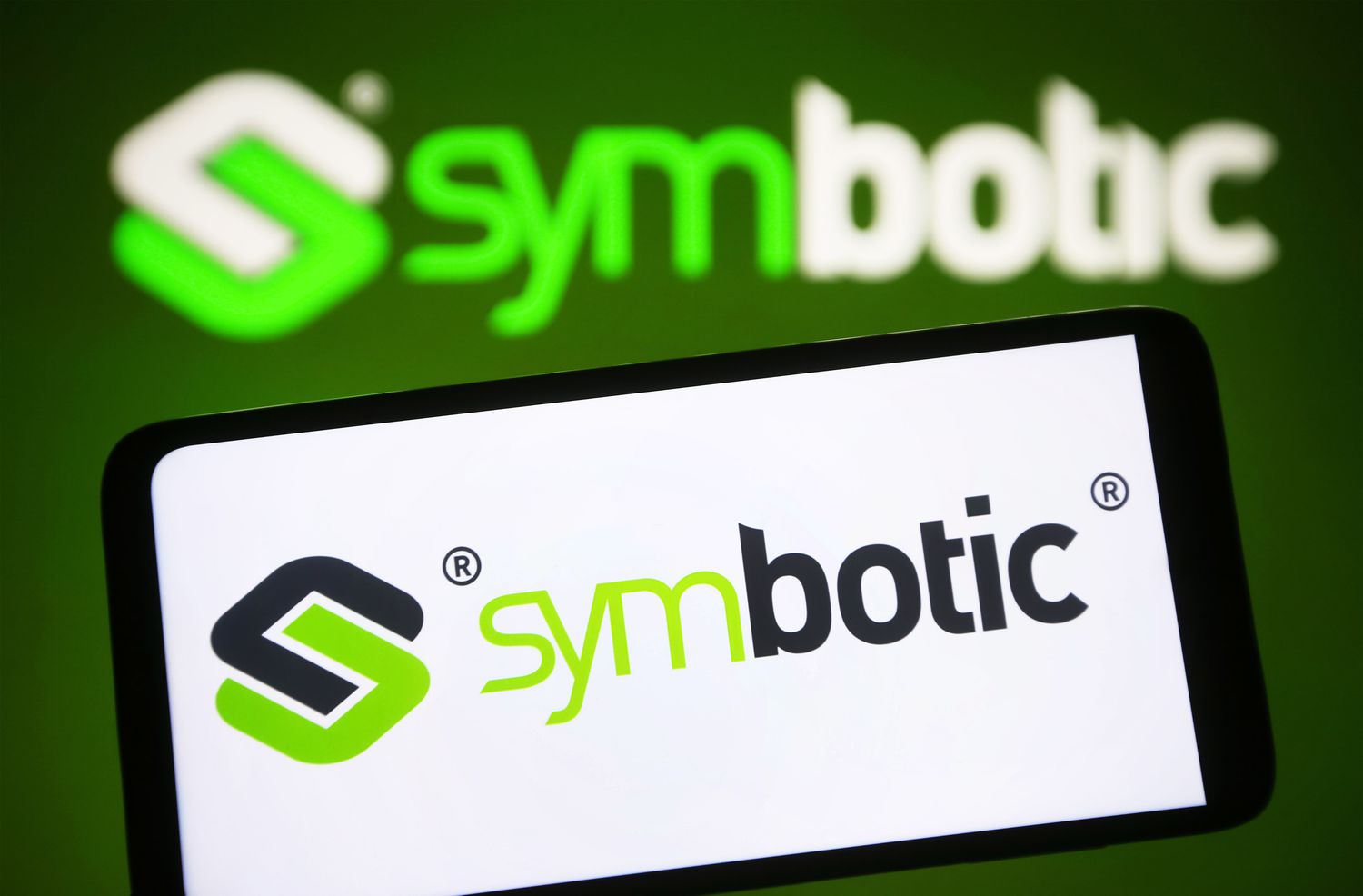 The green and black Symbotic logo is seen on a phone screen, with the logo also visible in the background.