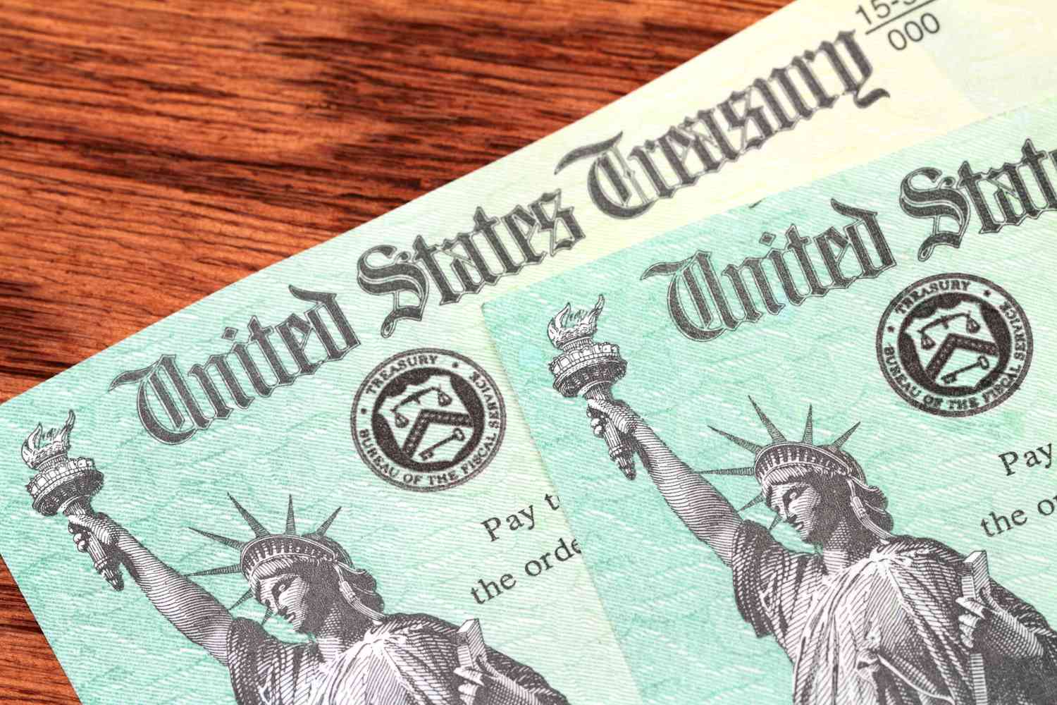 Two US Treasury checks of the type used to pay tax refunds as well as Medicare, Social Security and welfare benefits are partially visible on a wood desk background. Checks show the Treasury seal and image of the Statue of Liberty. 