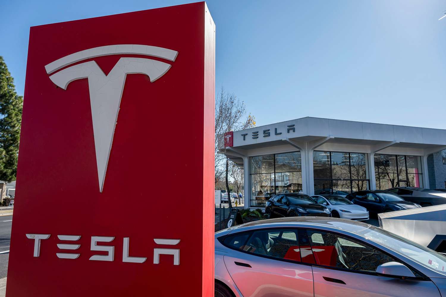 The Tesla logo is seen on a red sign with Tesla vehicles in front of a dealership building.