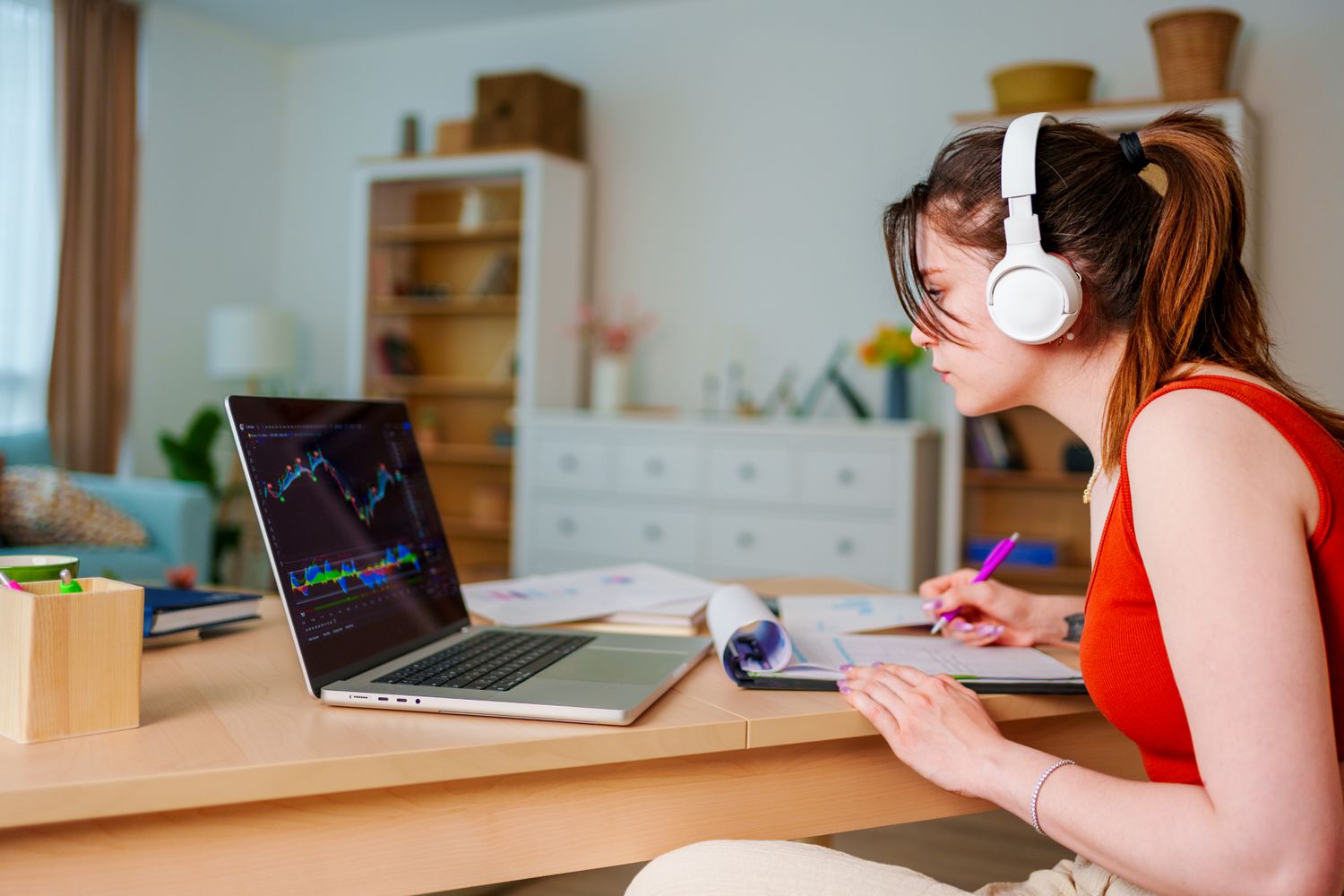 Young forex trader listening to the news while trading on laptop and also taking notes into trading journal. 