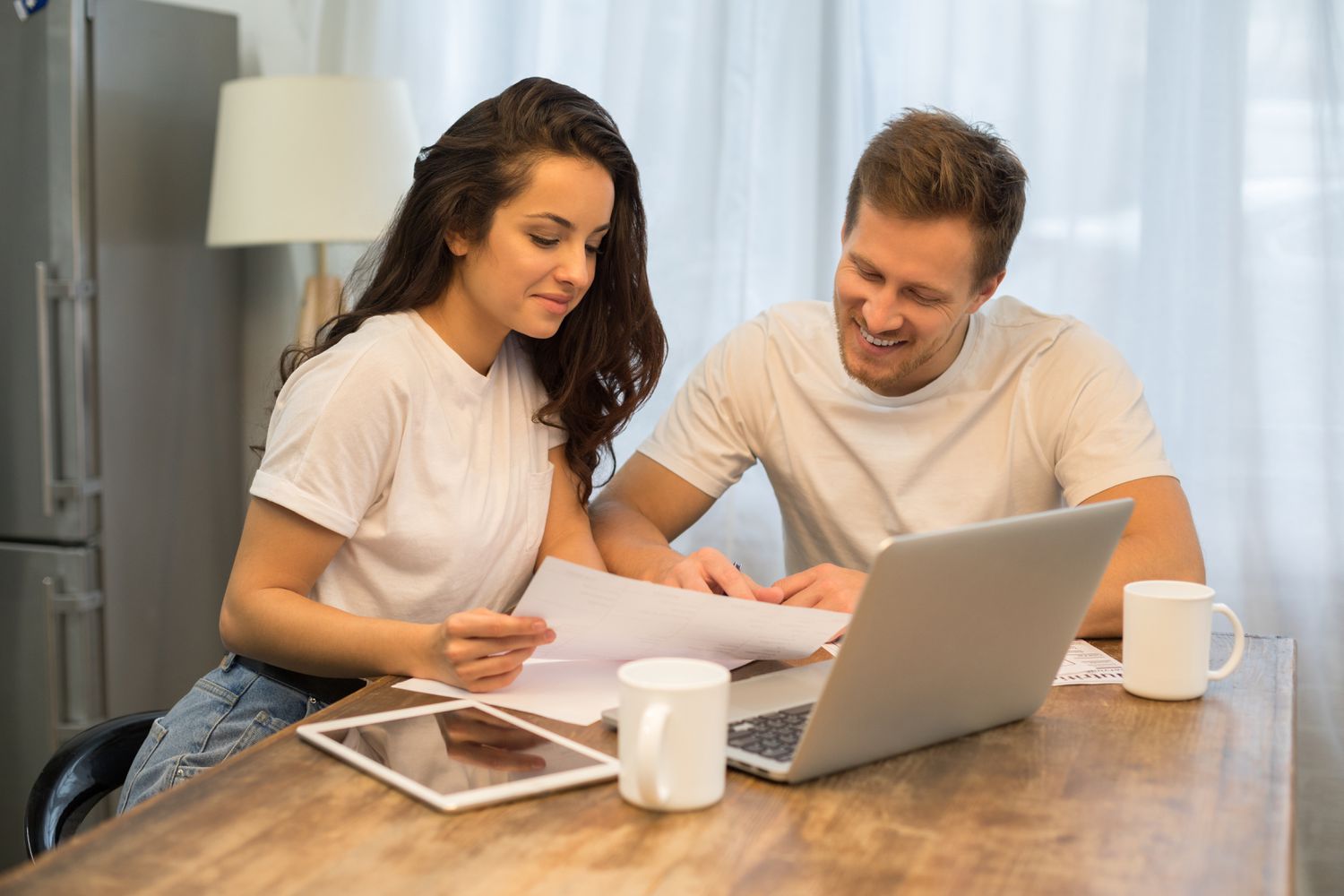 Young couple sitting at their kitchen table and looking happily at morgage documents and online rates.