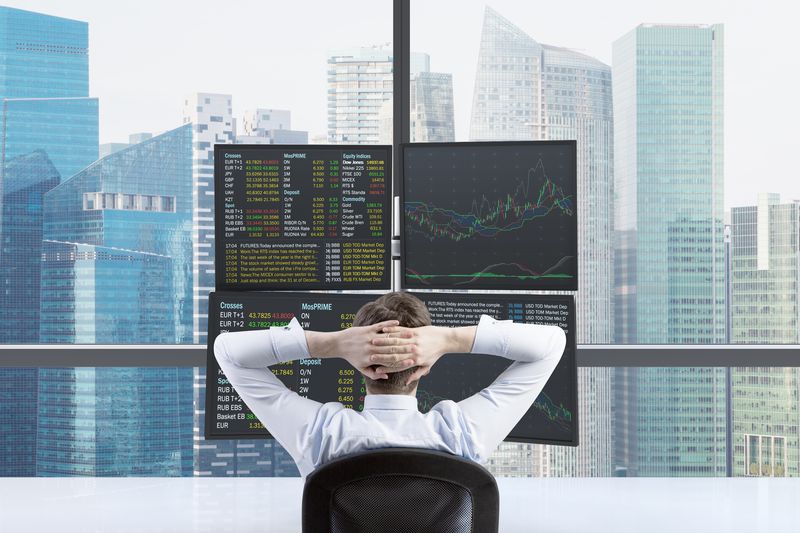 A forex arbitrage trader reclines in an office chair as they watch four monitors showing changes in international interest rates.