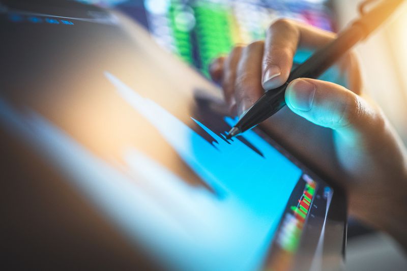 a hand holding a stylus used on a tablet with a chart with stock tickers in the background