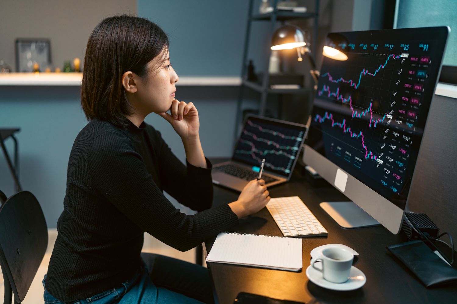 An investor looks at their home computer monitor and writes notes in a notebook about their trading strategy.