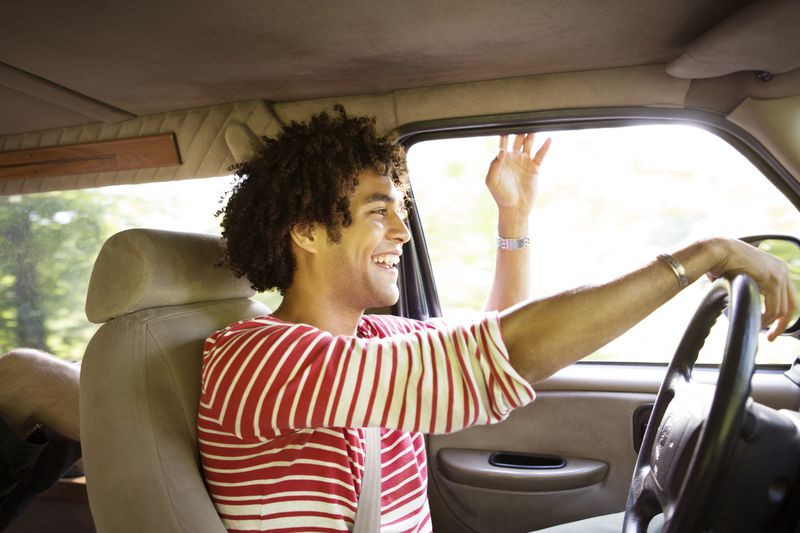 A happy, smiling man drives in a car with his hand out the window.