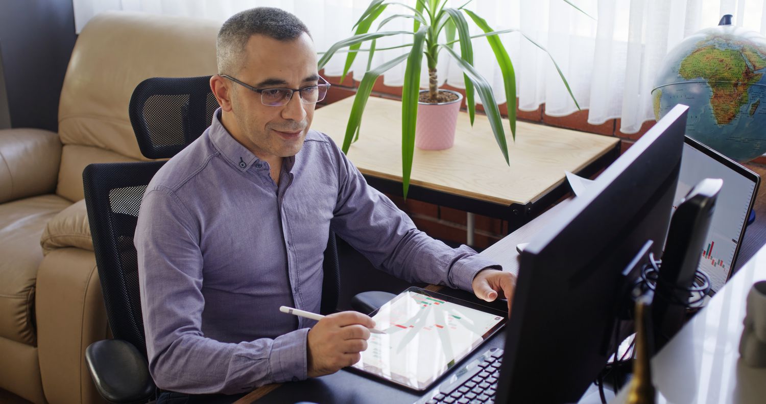 Man sitting at a desk looking at a computer monitor while making notes on a notepad.